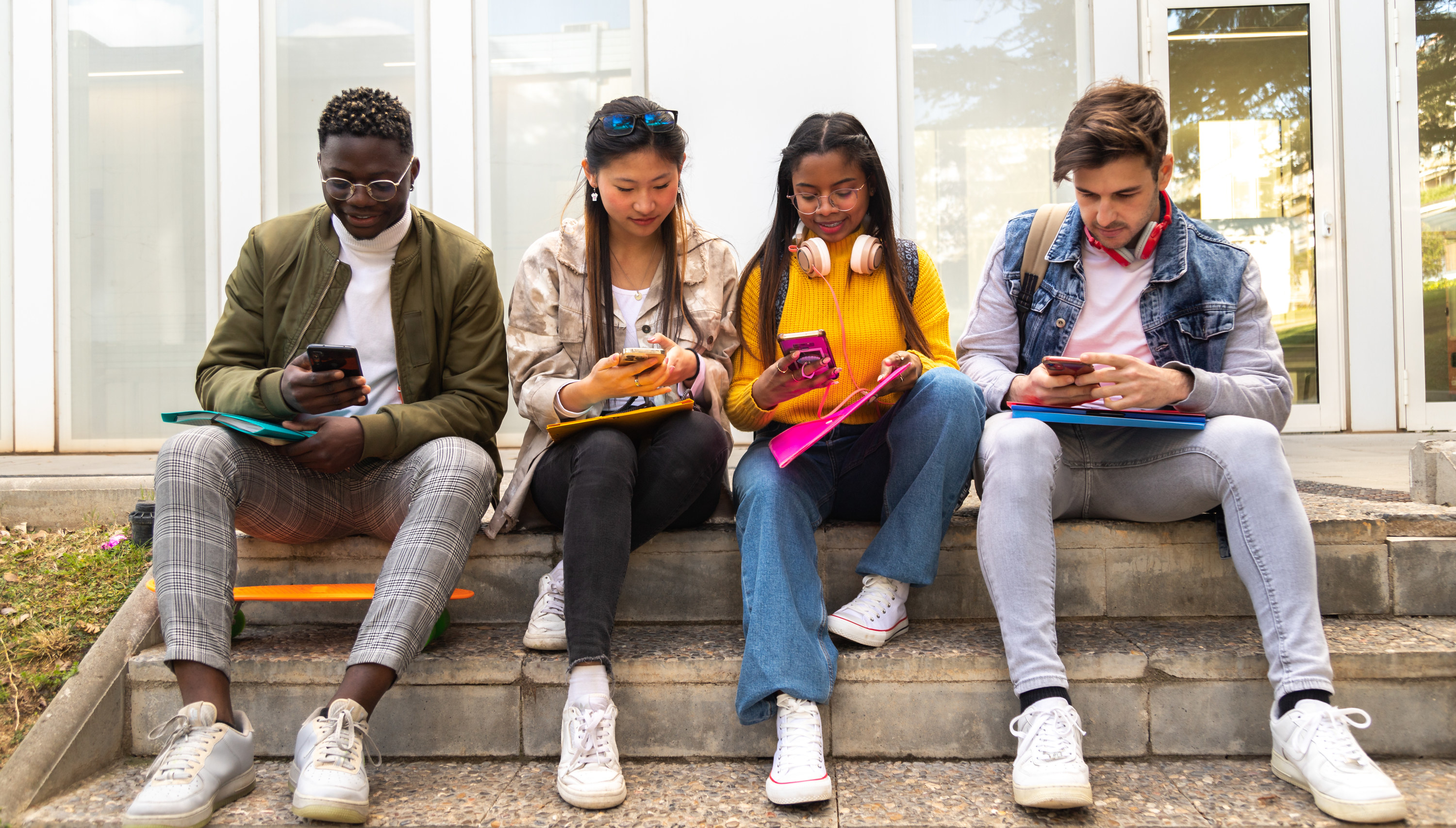 Four students use their phones while sitting next to one another outside