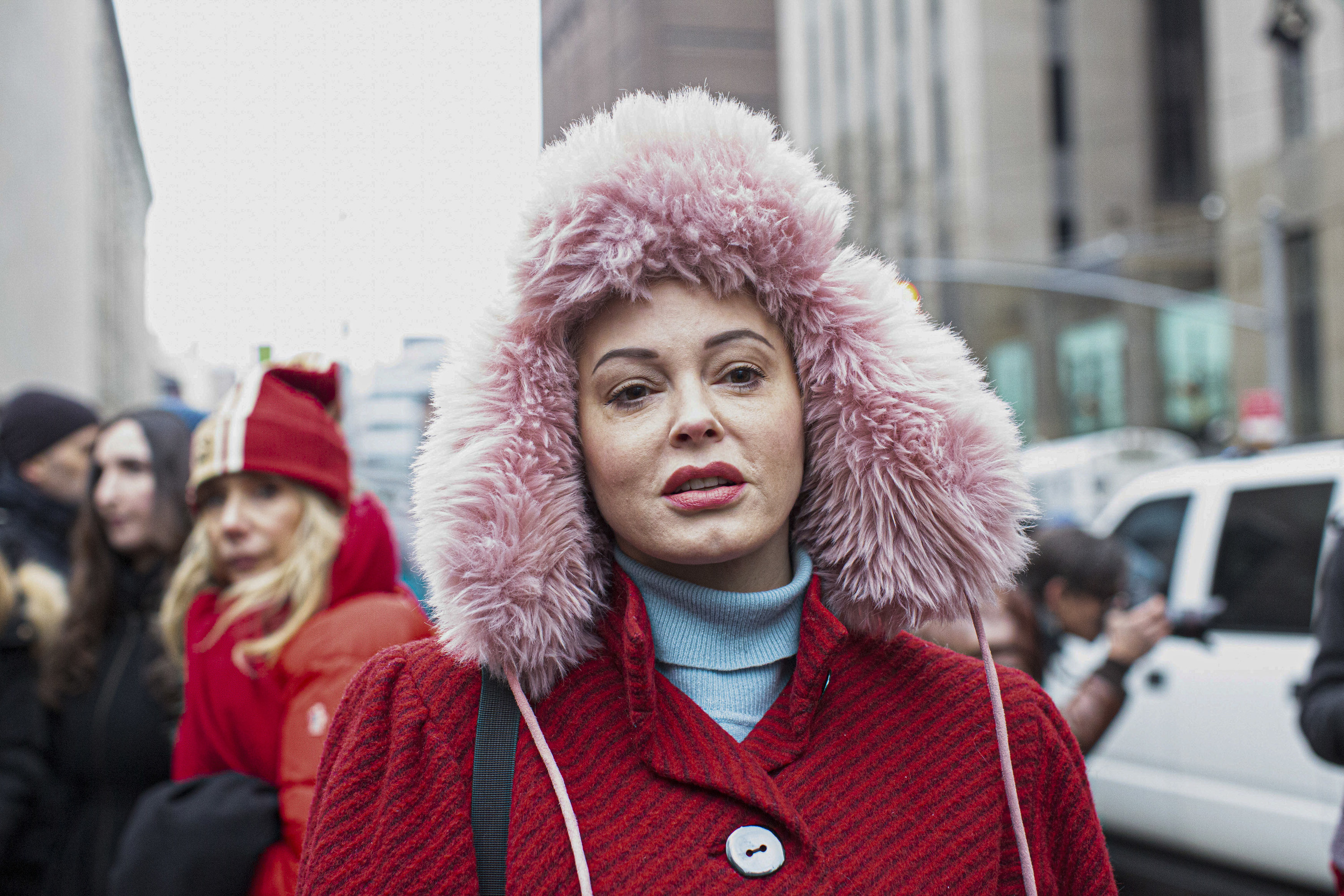 Rose McGowan walks in a crowd in a red coat and big pink fluffy hat