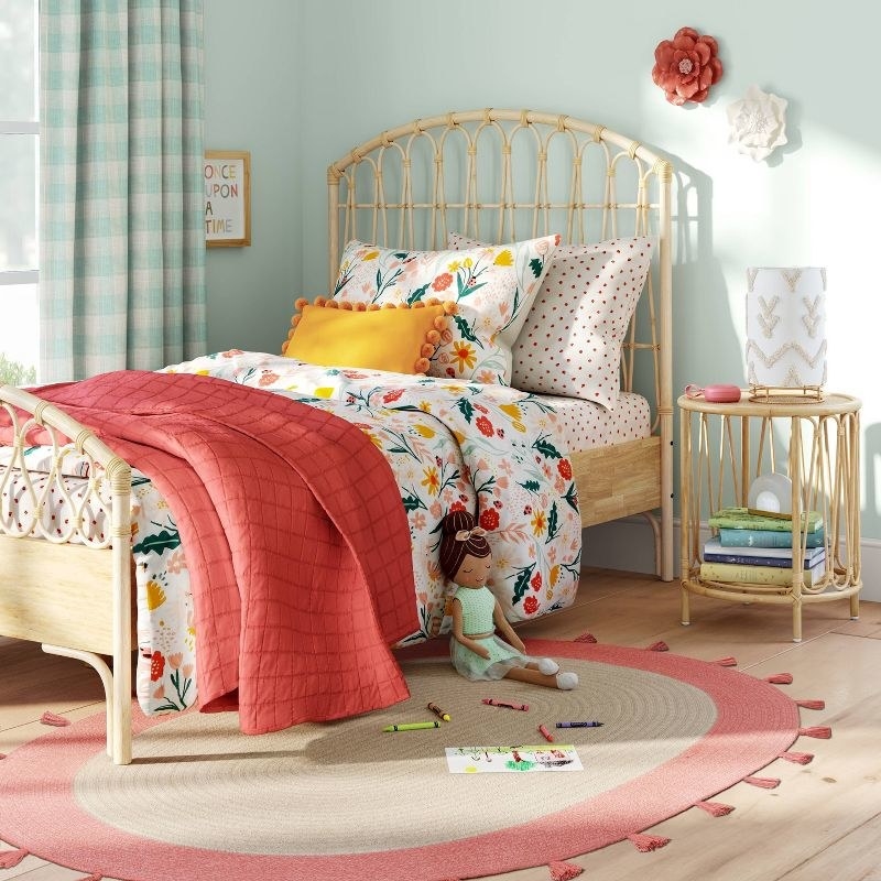 a child&#x27;s bedroom with a made bed, an oval rug, a doll, and a rattan bedside table