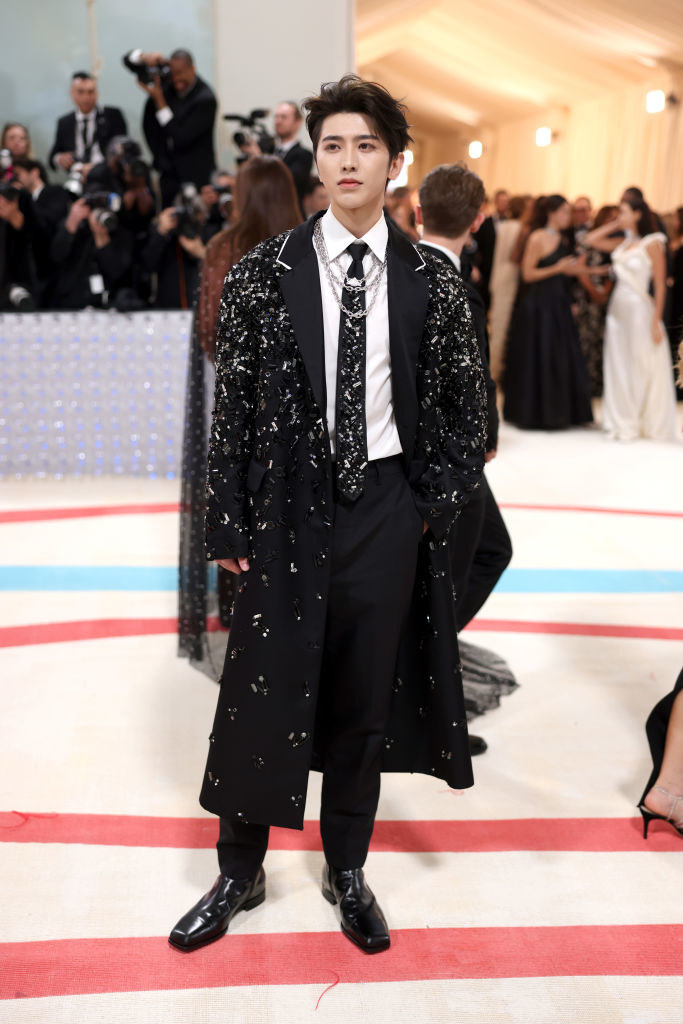 in a long coat with sparkled detailing and diamond necklaces layered with his tie