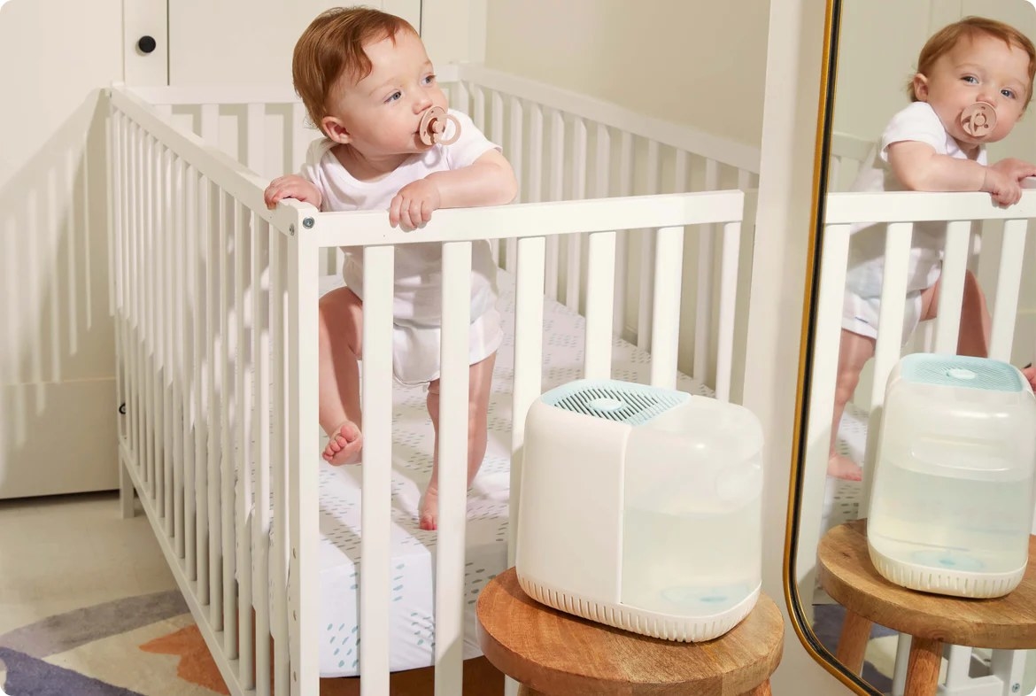 Child model in crib with white and blue humidifier on nearby bench