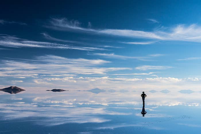 clear blue sky and a person standing in the middle of the salt flat with the clouds reflected below their feet
