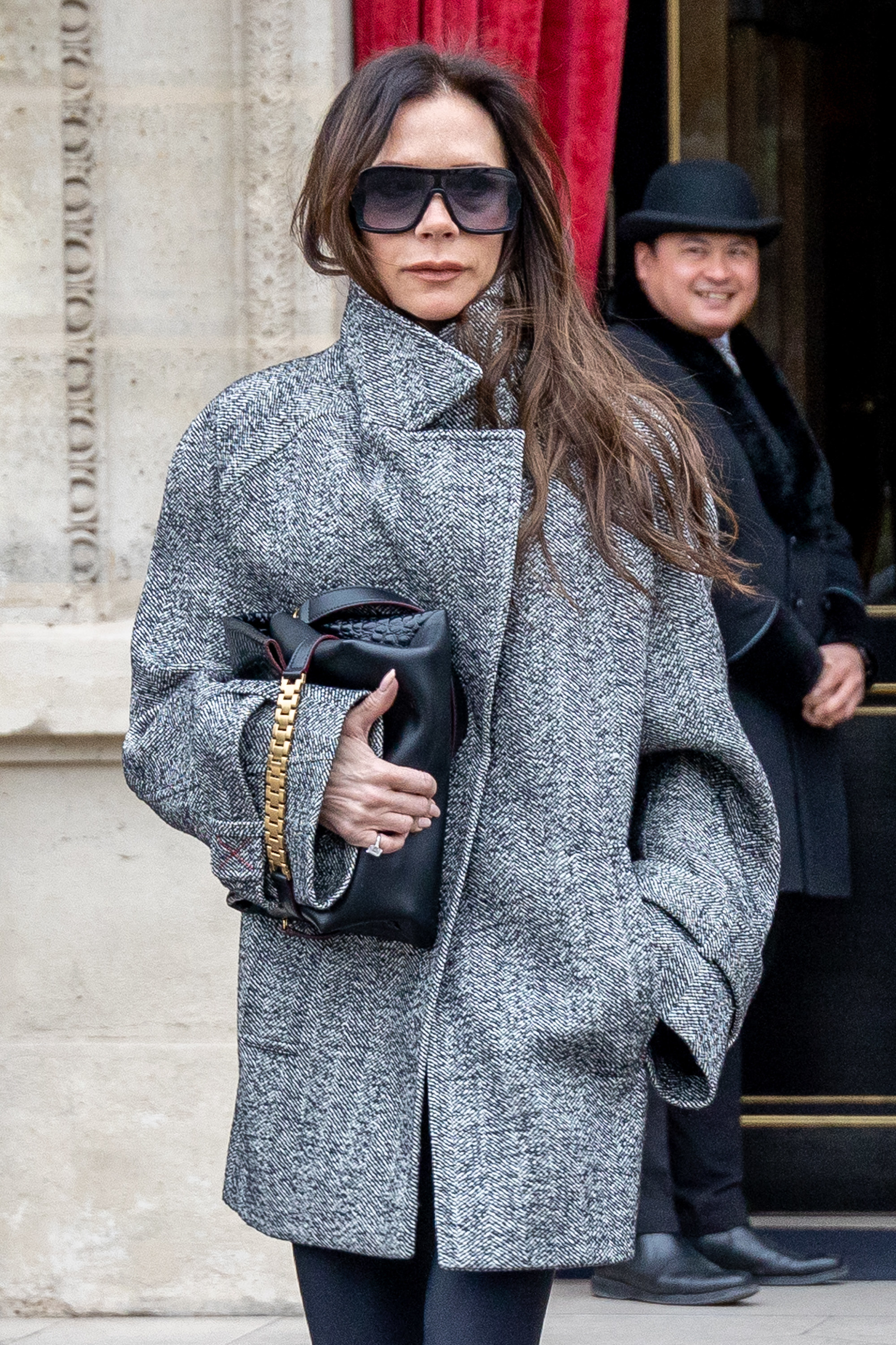 Victoria Beckham, wearing a big grey coat and oversized sunglasses, walking out of a building