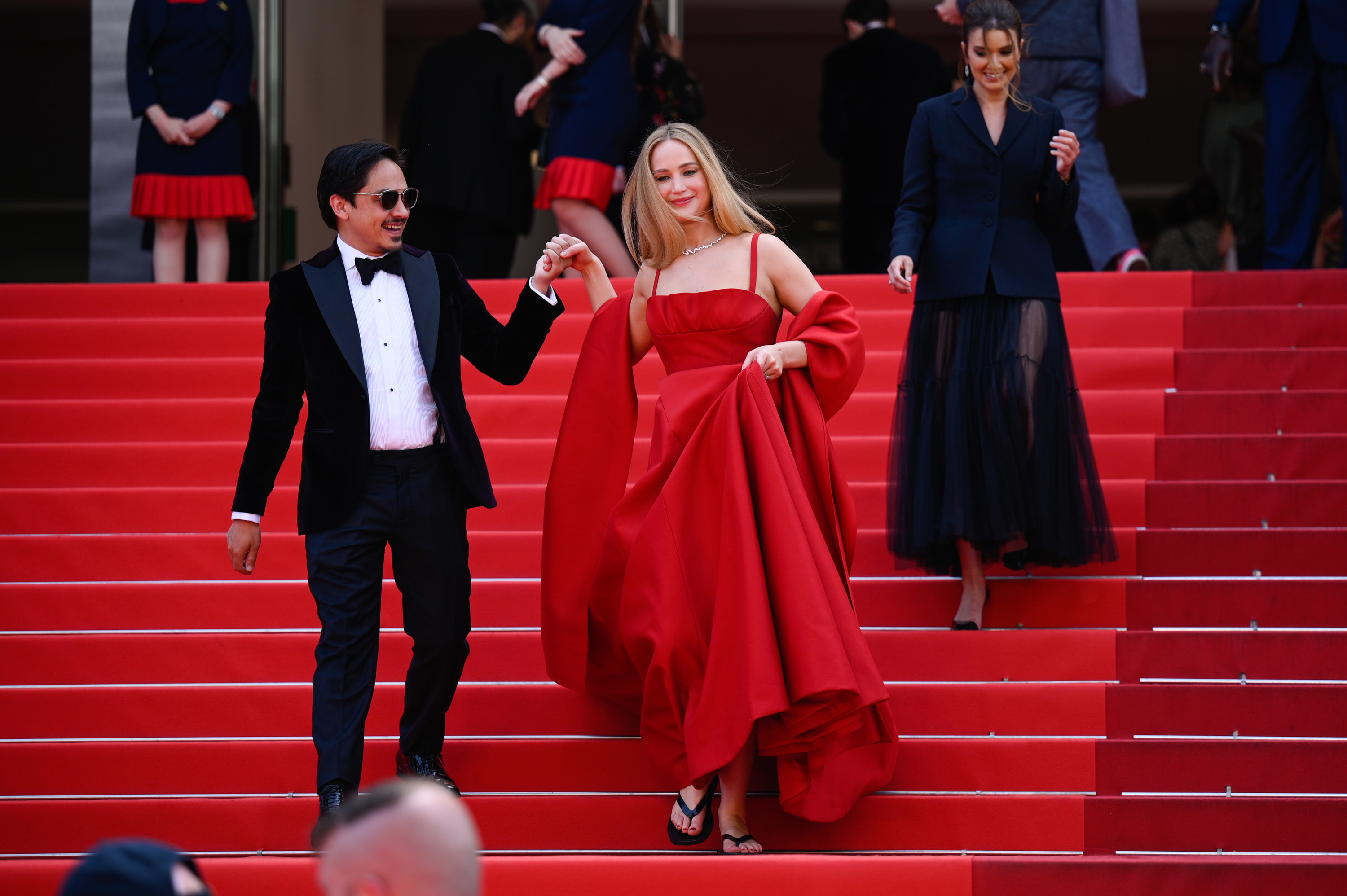 JLaw on the red carpet stairs raising her spaghetti-strap red gown to show black flip-flops