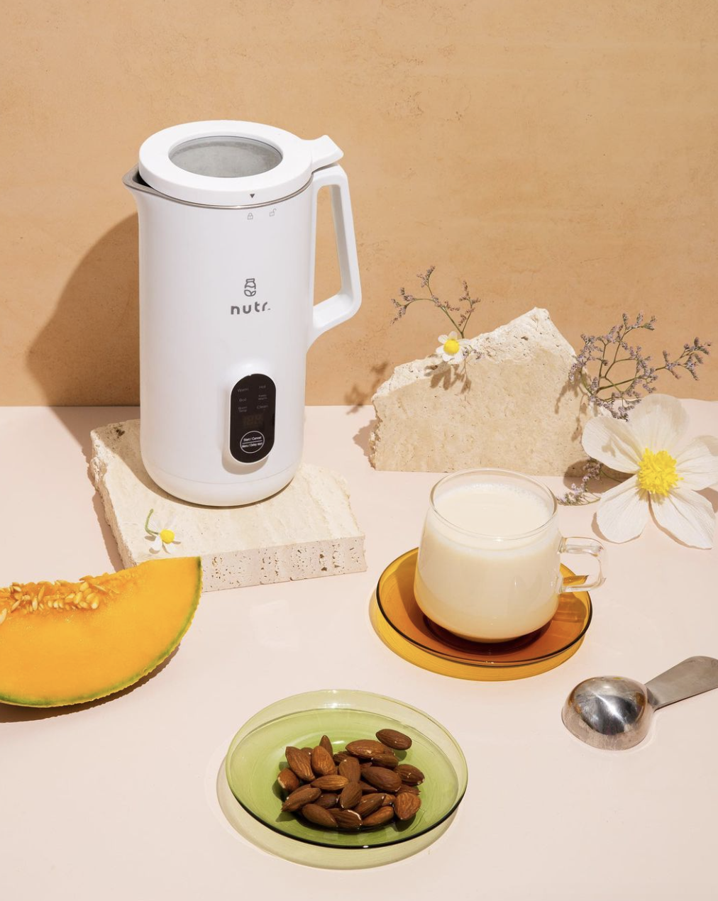 nut milk machine beside latte and plate of almonds with cantaloupe and flowers nearby