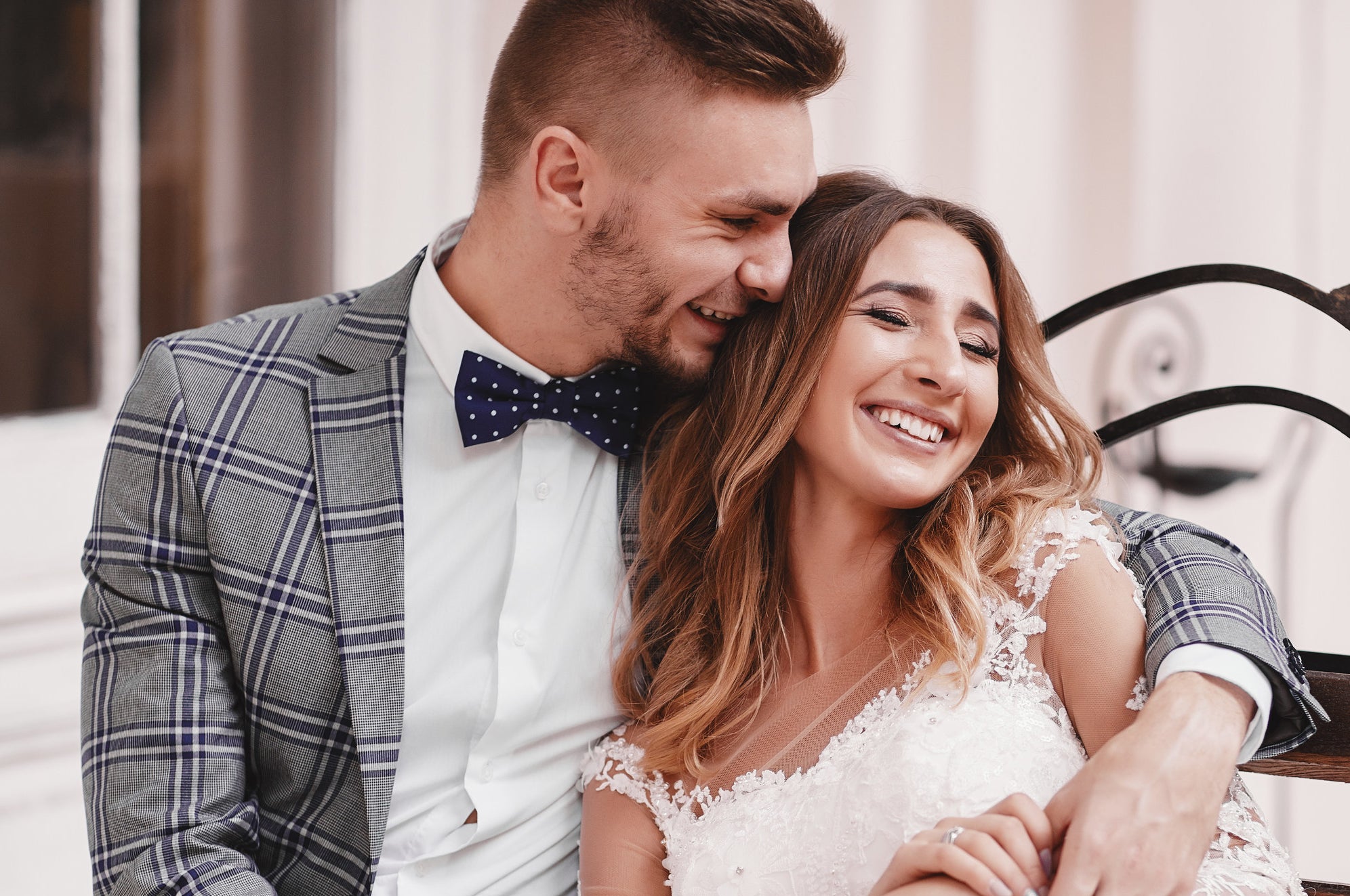 A groom and bride smile and embrace while sitting on a bench