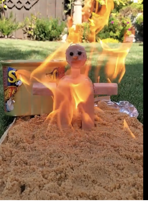 A &quot;Burning Man&quot;-type figure on fire above a sheet of Spam lying on the grass