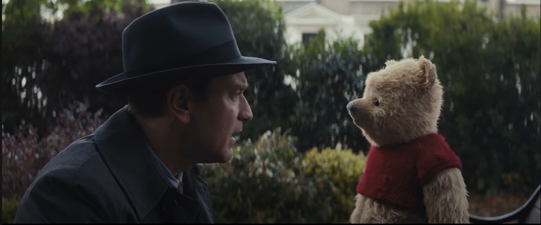 Man in a hat sitting beside an animated bear in a red sweater, both appear engaged in conversation