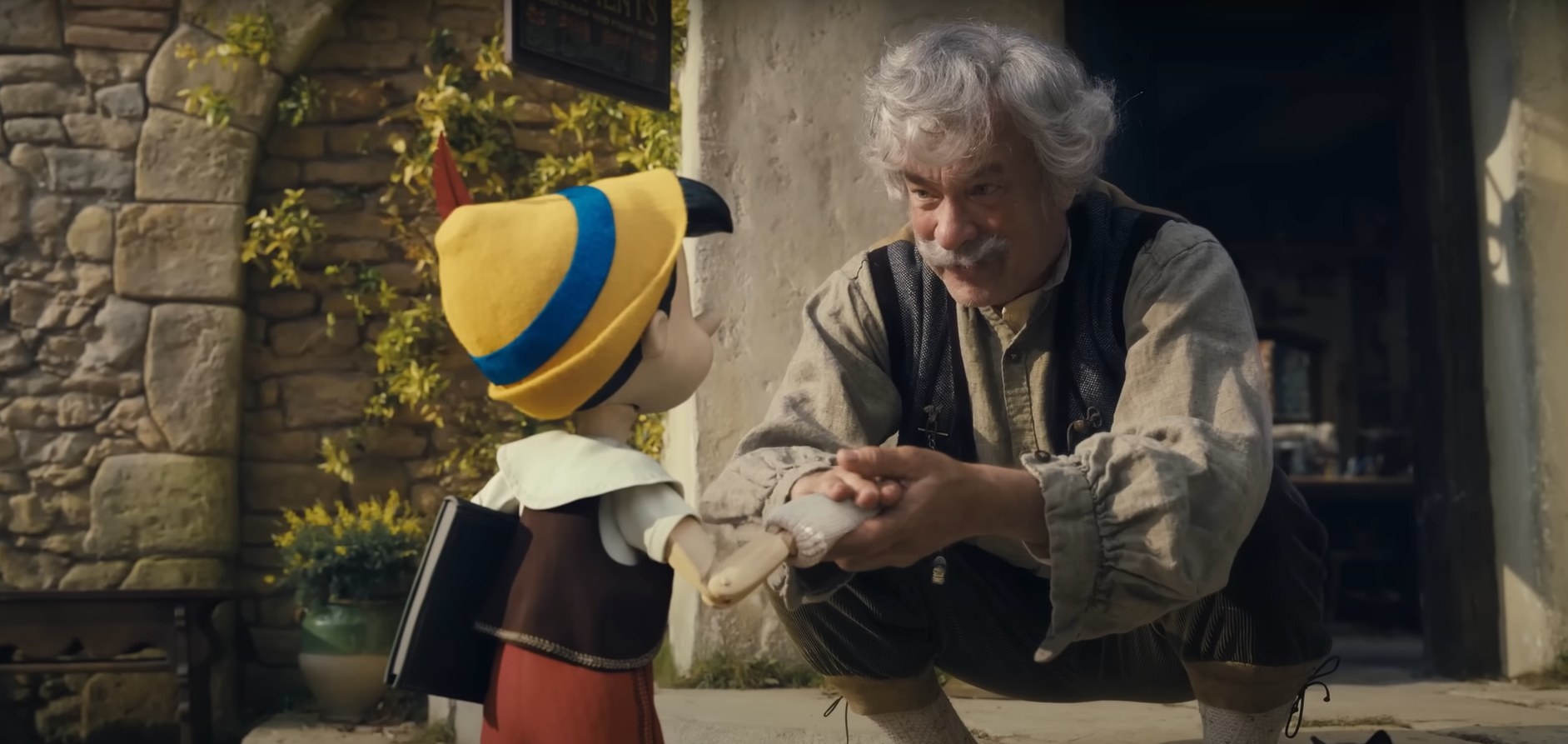 Geppetto, a live-action character, kneels to talk to Pinocchio, an animated figure. They are exchanging a heartfelt gesture