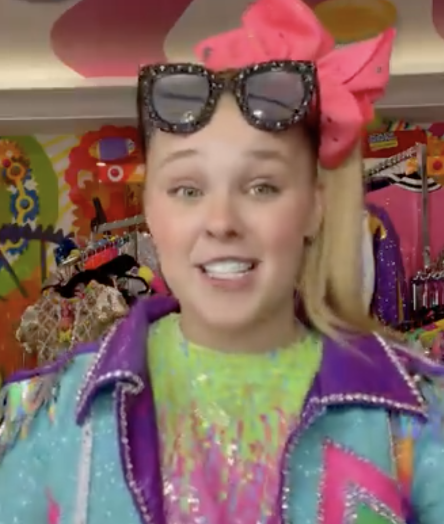 A screenshot of JoJo Siwa standing in front of the camera, with a sequined multicolored jacket and shirt, huge black-and-diamond sunglasses, and a giant pink bow