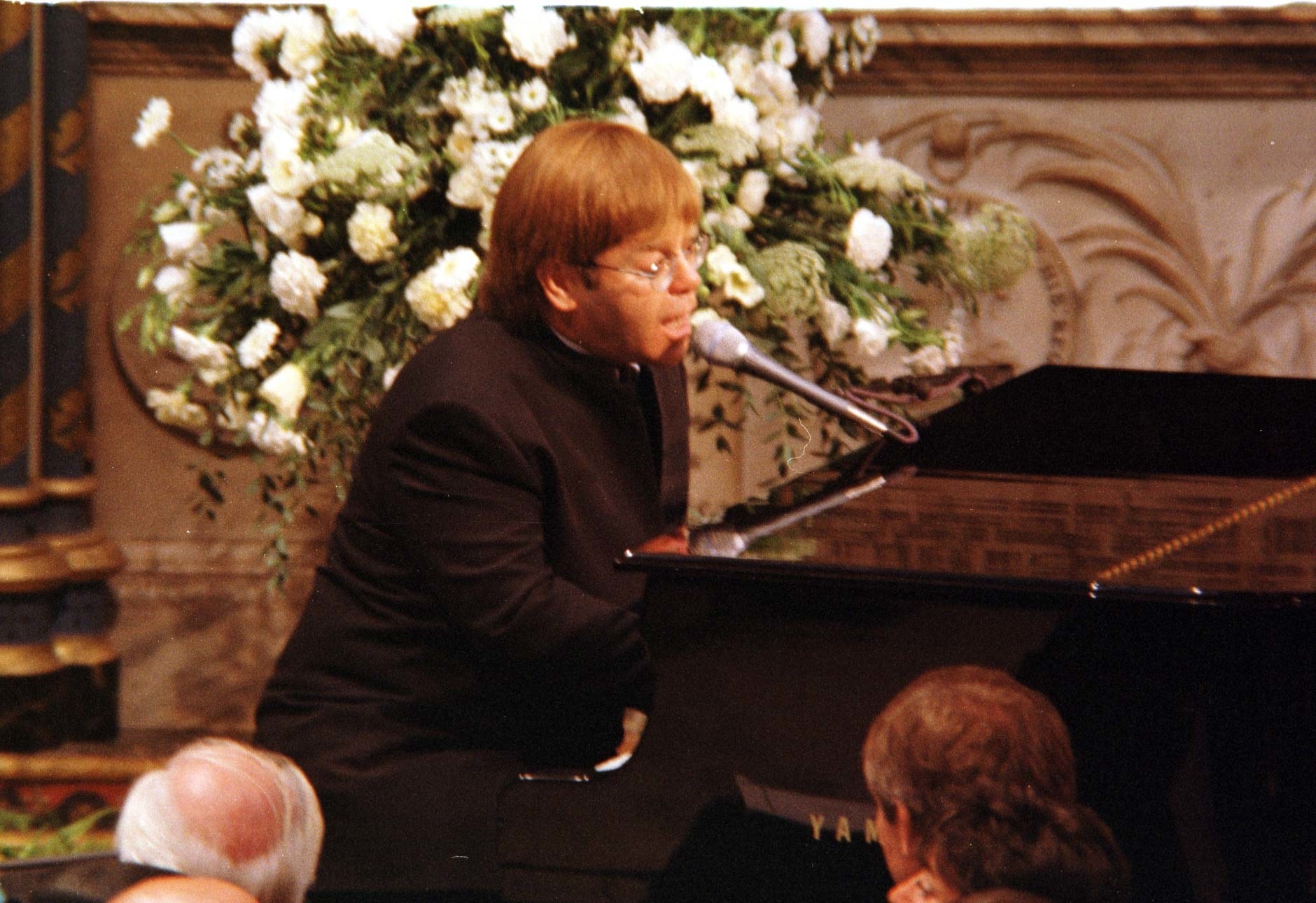Elton John sitting at a piano and singing, wearing black, with a church altar behind him