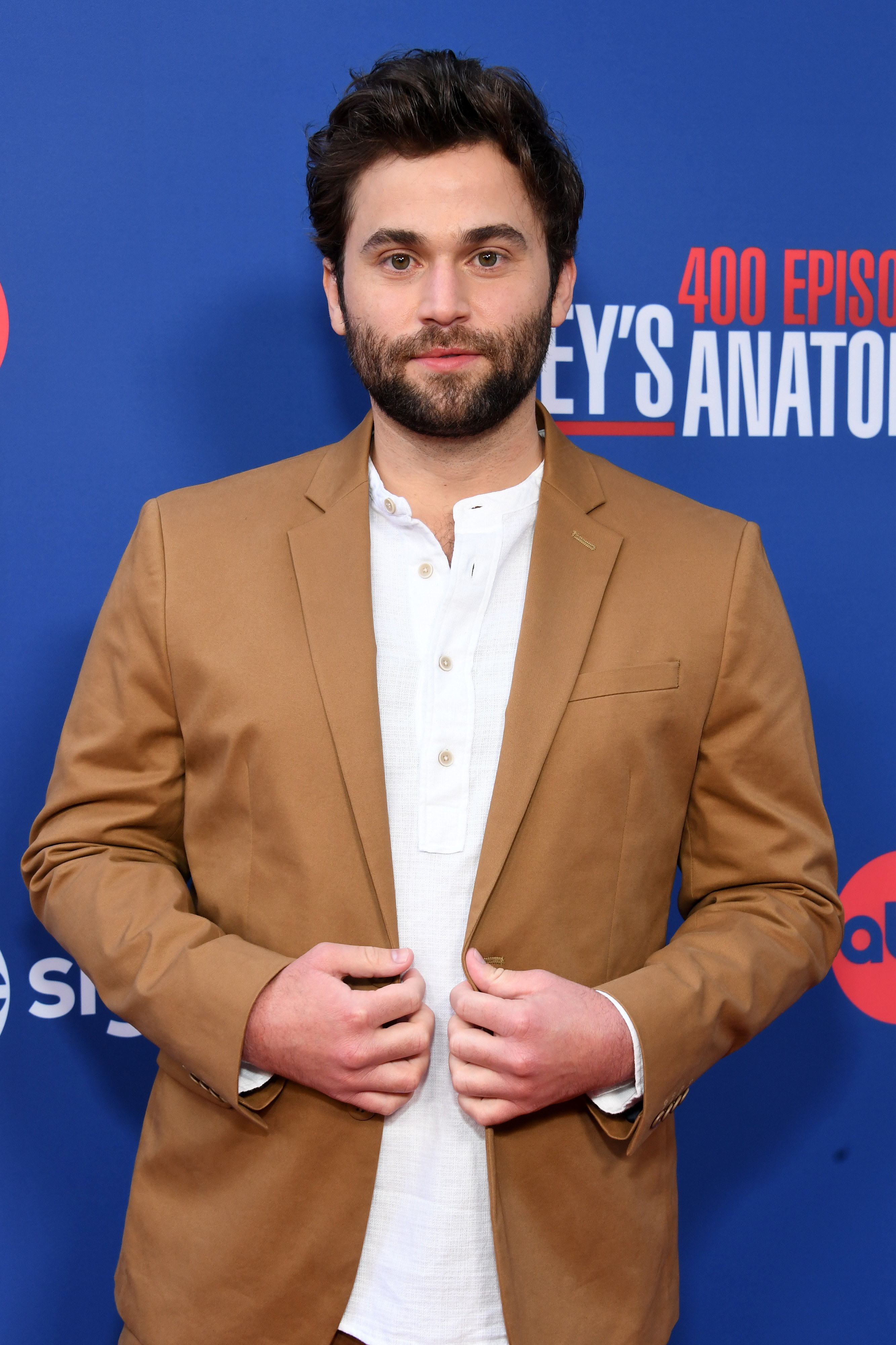 Jake Borelli posing at a Grey&#x27;s Anatomy event, wearing a white linen shirt and a tan blazer