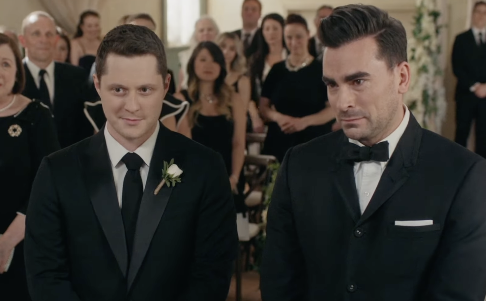David and Patrick from Schitt&#x27;s Creek, wearing black suits and standing at the front of a room with a crowd of people standing behind them