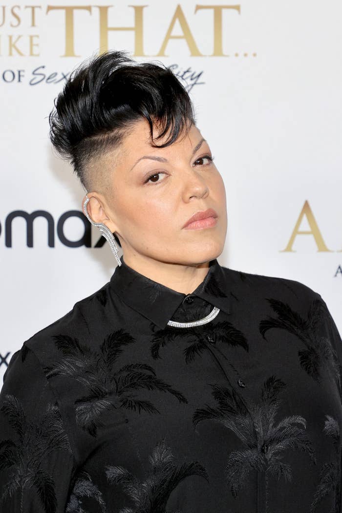 Sara Ramirez poses on the red carpet in a buttoned up black shirt pattered with silk palm trees, and a thick silver chain around their neck