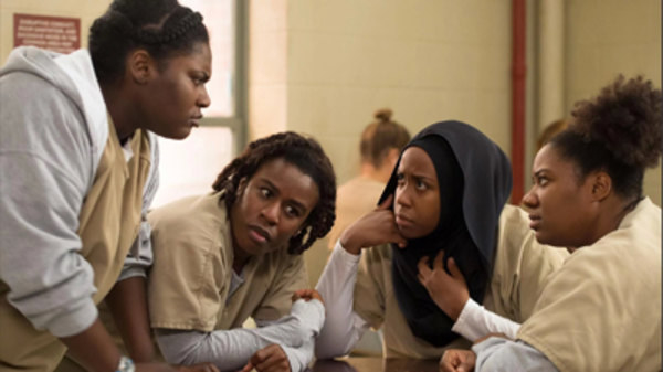 A screenshot from OITNB, showing four characters wearing tan scrubs sitting at a table