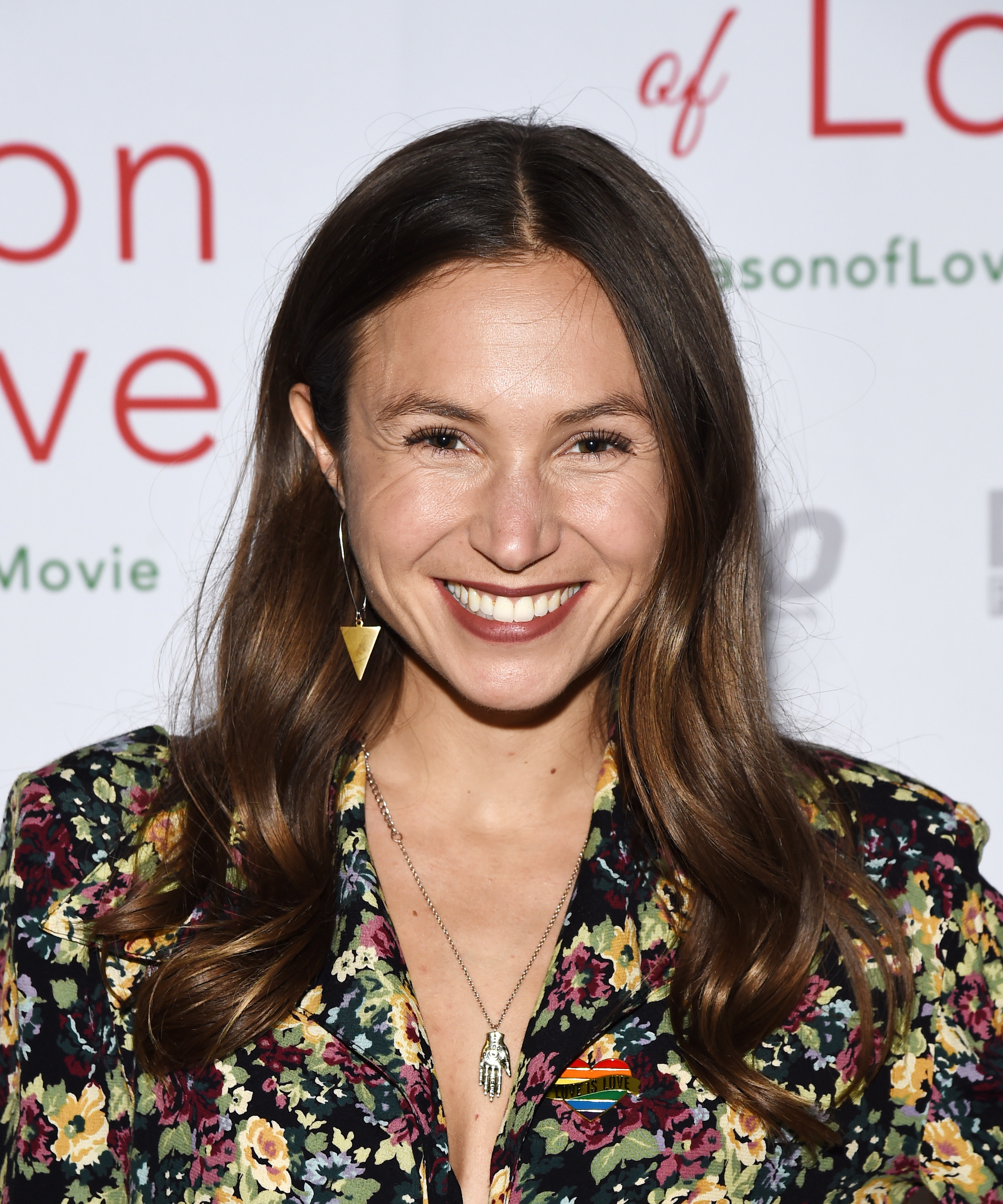 Dominique Provost-Chalkley posing at an event wearing a flowery top that has a rainbow heart pin on it
