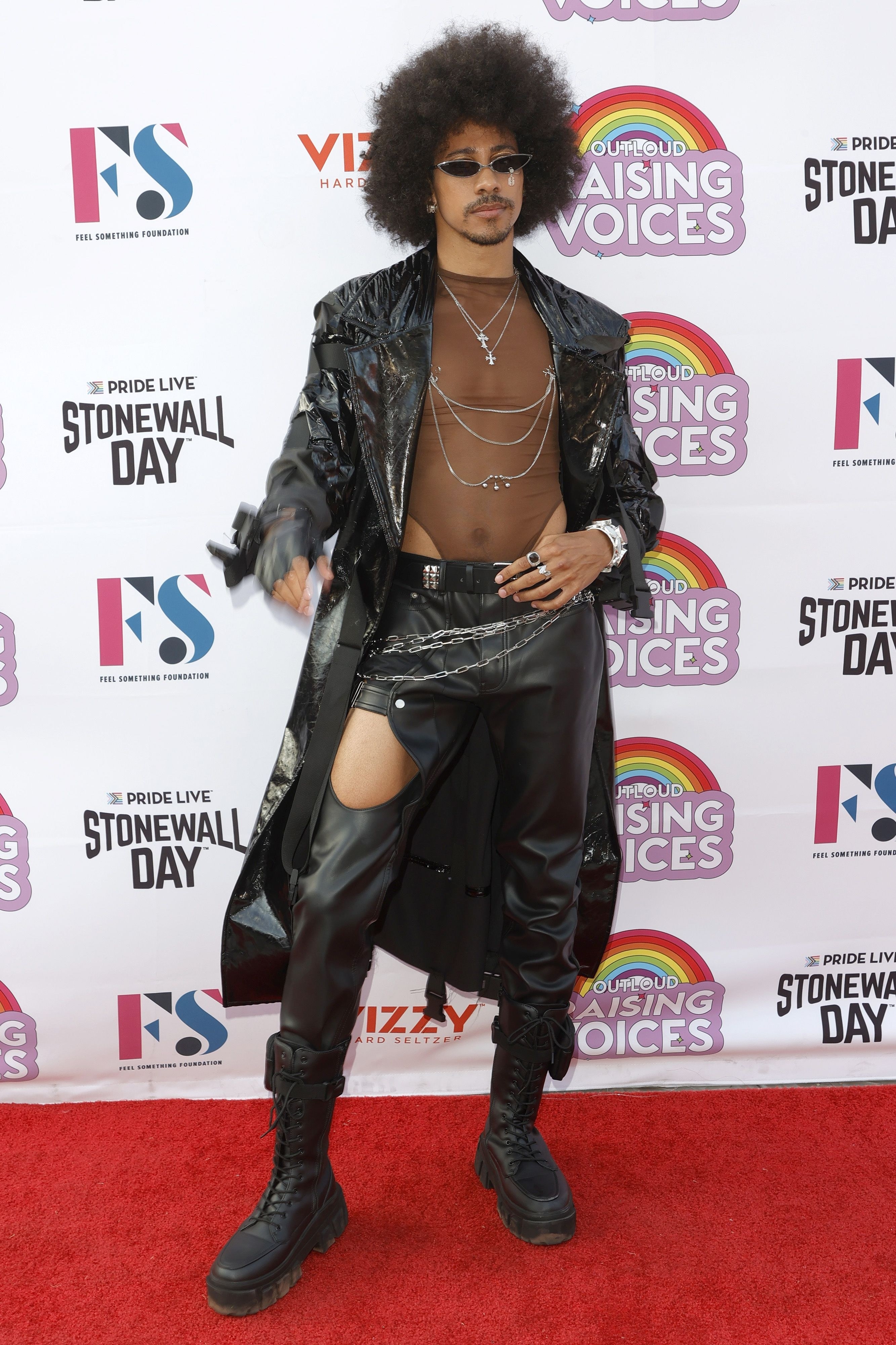 Keiynan Lonsdale poses on the red carpet in an all-black outfit with boots, leather pants, a mesh bodysuit, and long black coat, with sunglasses