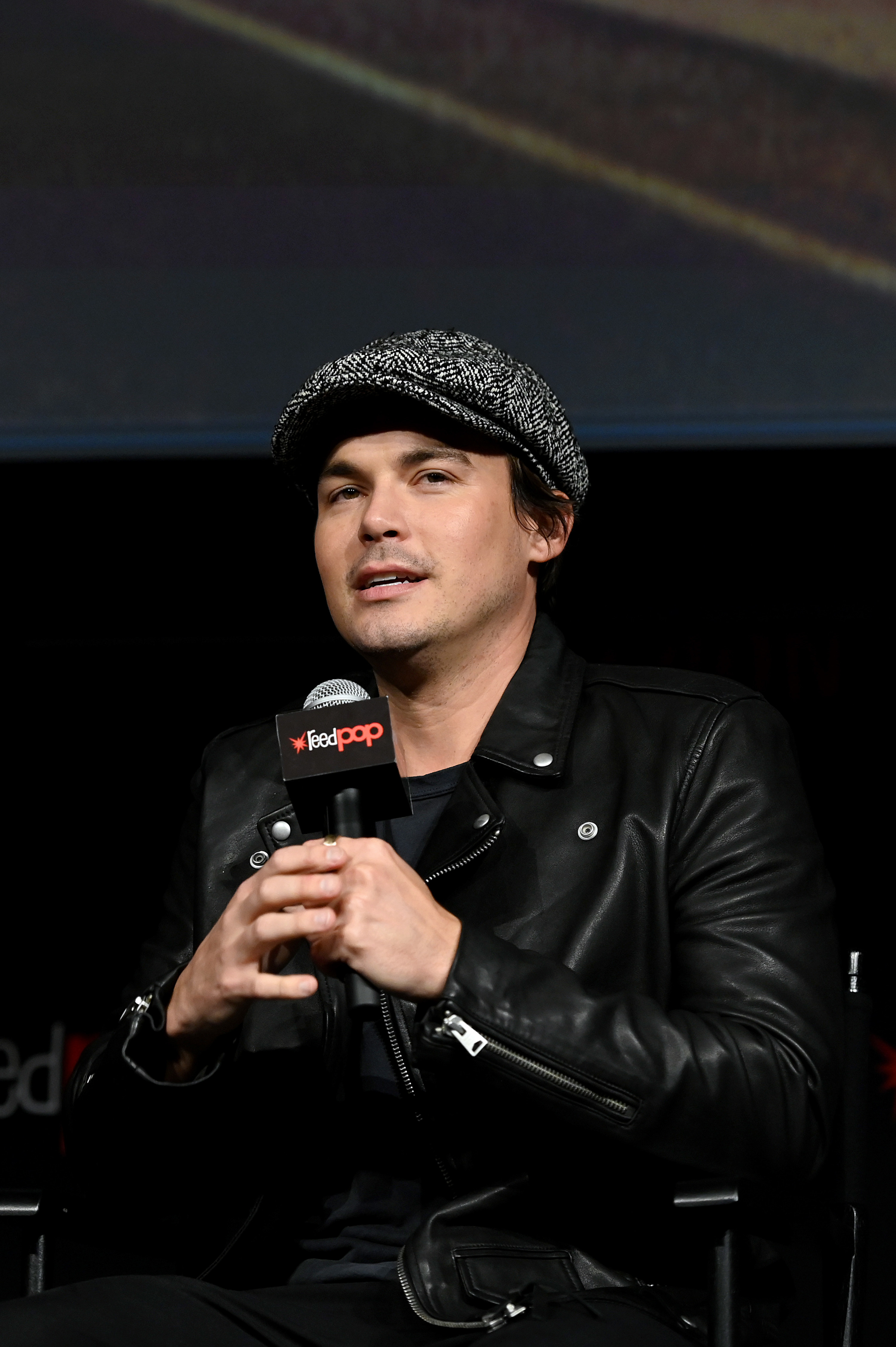 Tyler Blackburn holding a microphone and sitting, wearing a tweed cap and leather jacket