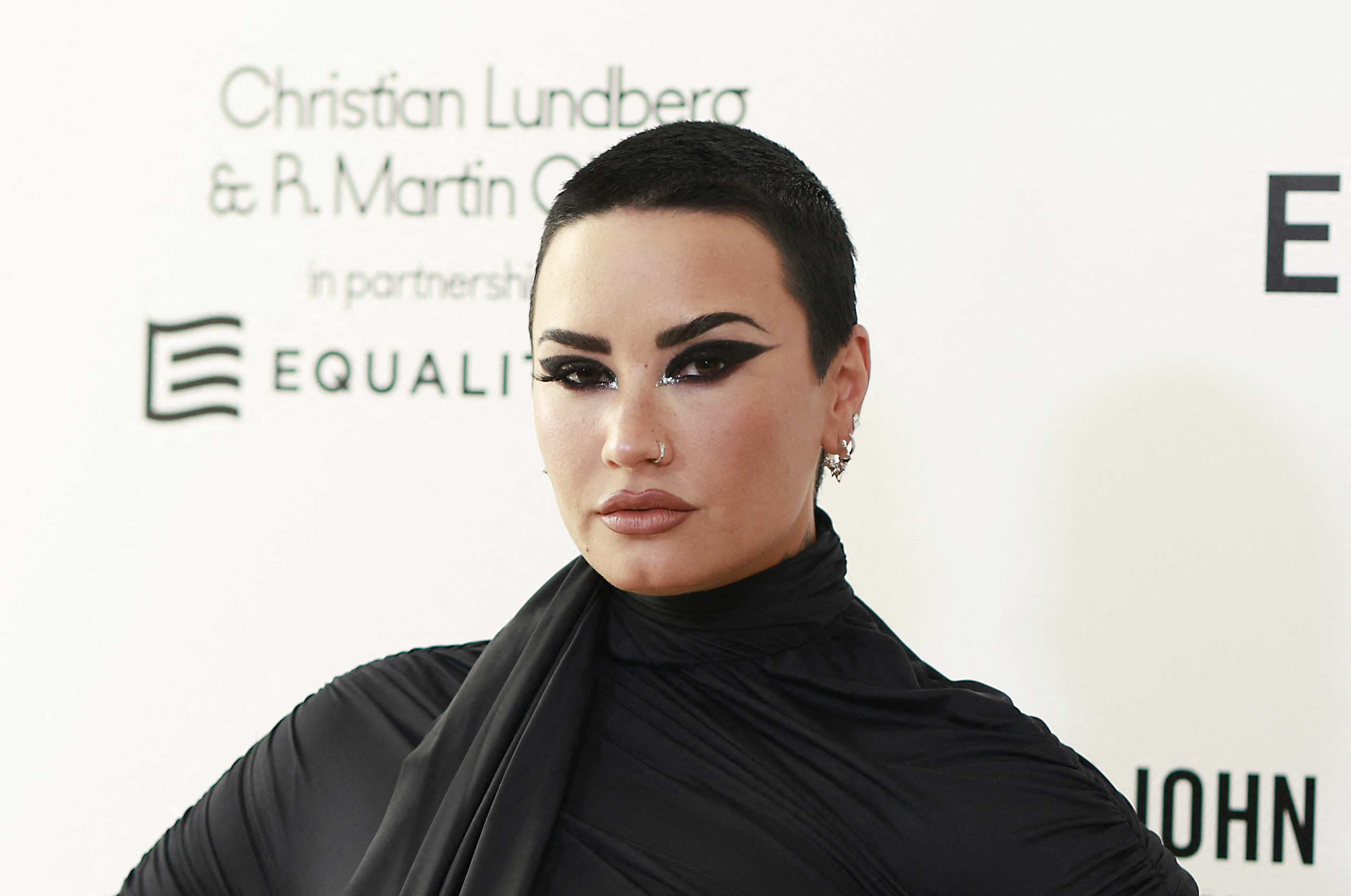 Demi Lovato posing at an event with a short black buzzcut, dark eyeliner, and a black turtleneck