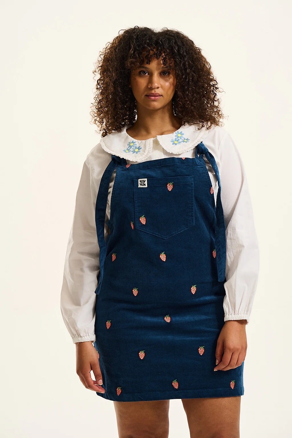 model in dark blue corduroy mini overall dress with tie straps and an embroidered strawberry print pattern