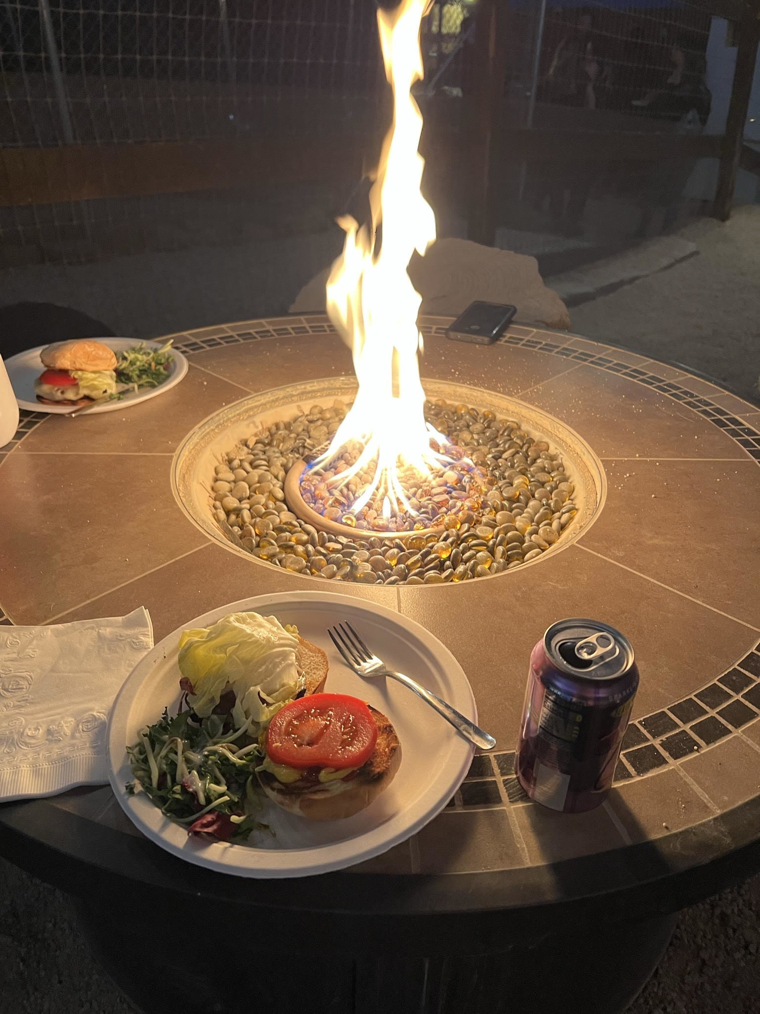 A fire in the center of a table outdoors and plates of food and beverages