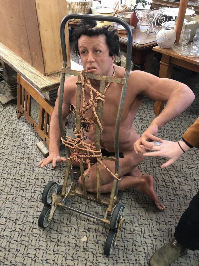 A life-size naked Sylvester Stallone doll on its knees and attached to a cart with rope