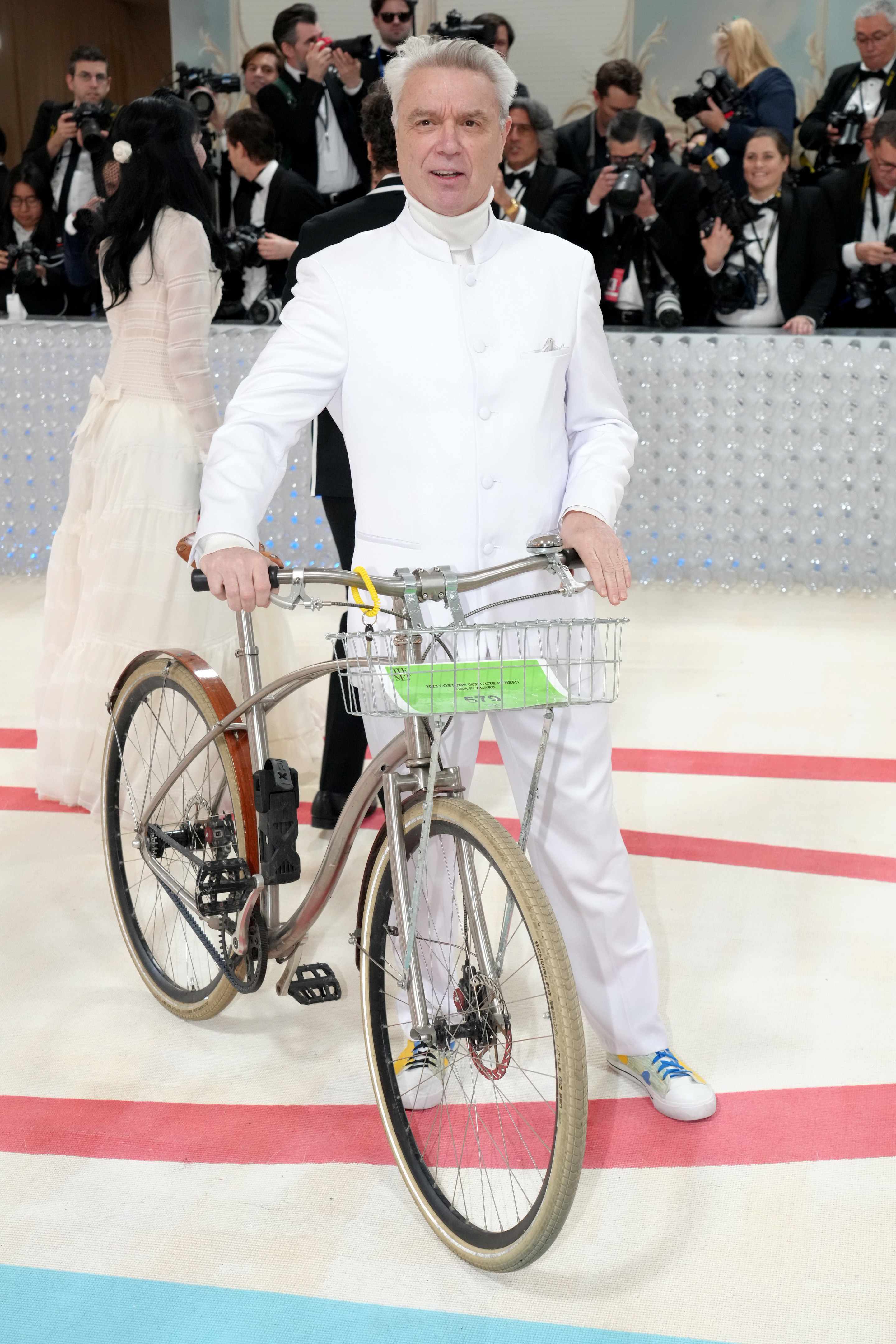 David Byrne wearing an all-white suite, sneakers, and holding a bike on the red carpet