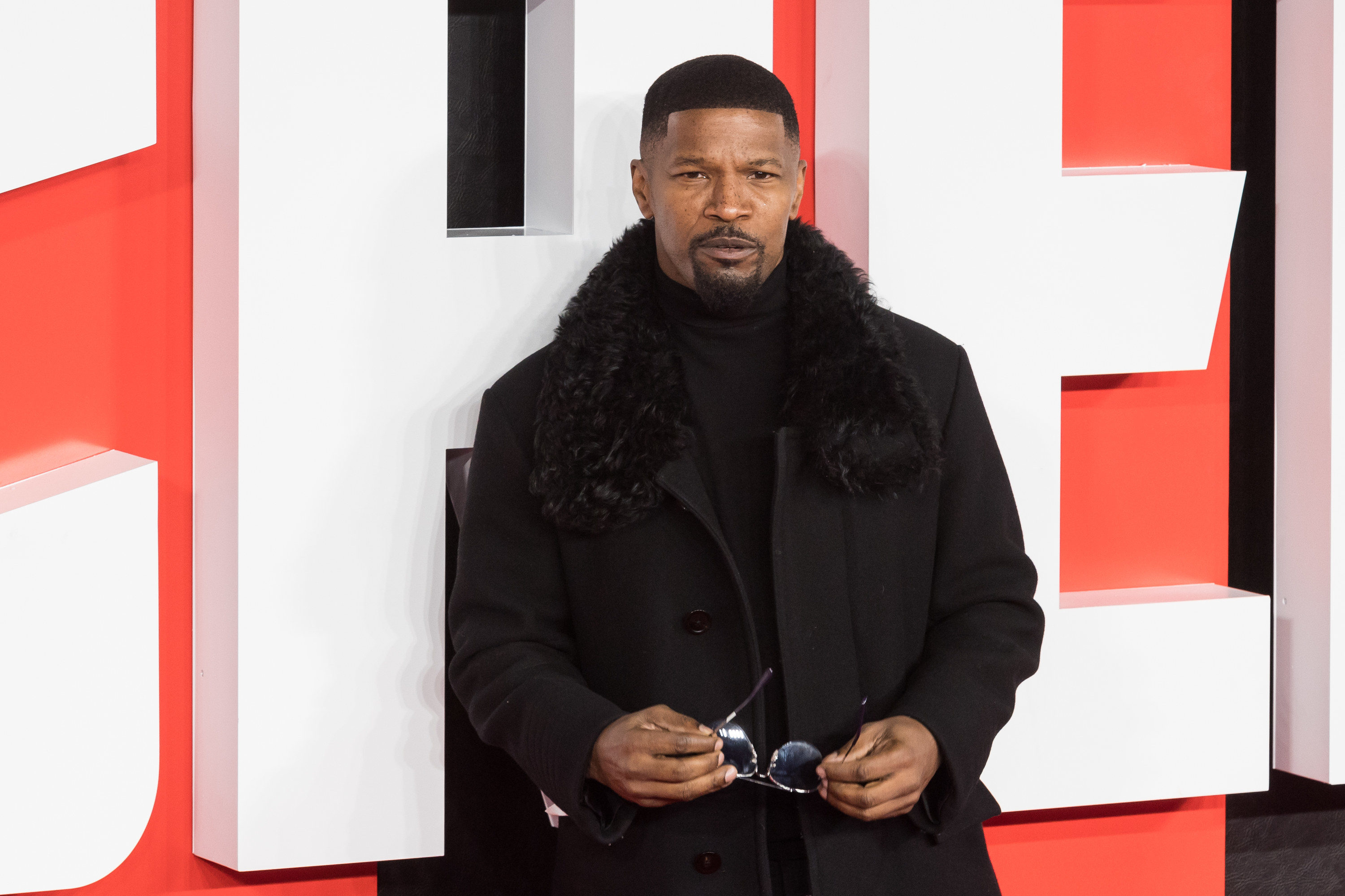 Closeup of Jamie Foxx in a fitted coat and holding a pair of sunglasses on the red carpet