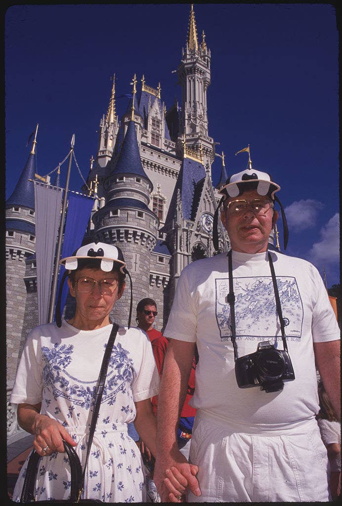 two adults, both wearing Goofy the character hats, holding hands in front of the disney castle