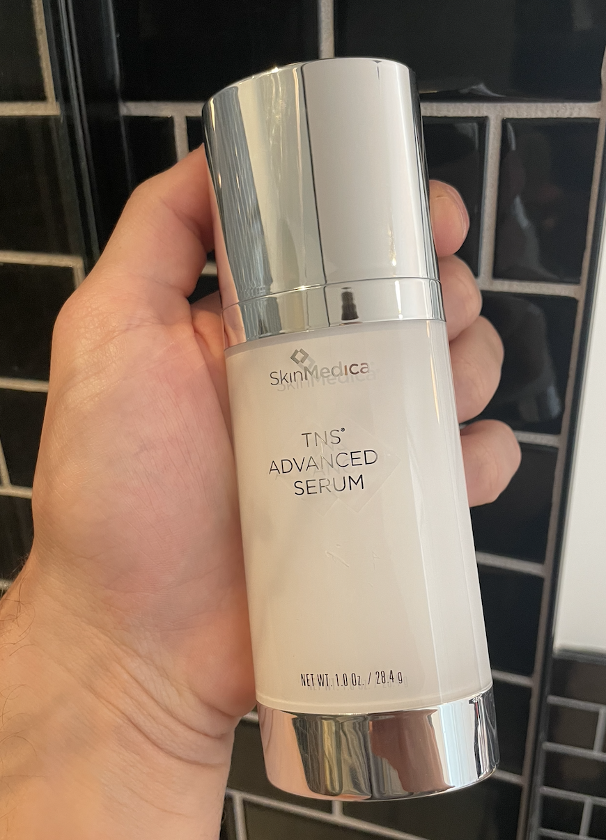 a man holds a bottle of large bottle SkinMedica TNS Serum in a bathroom