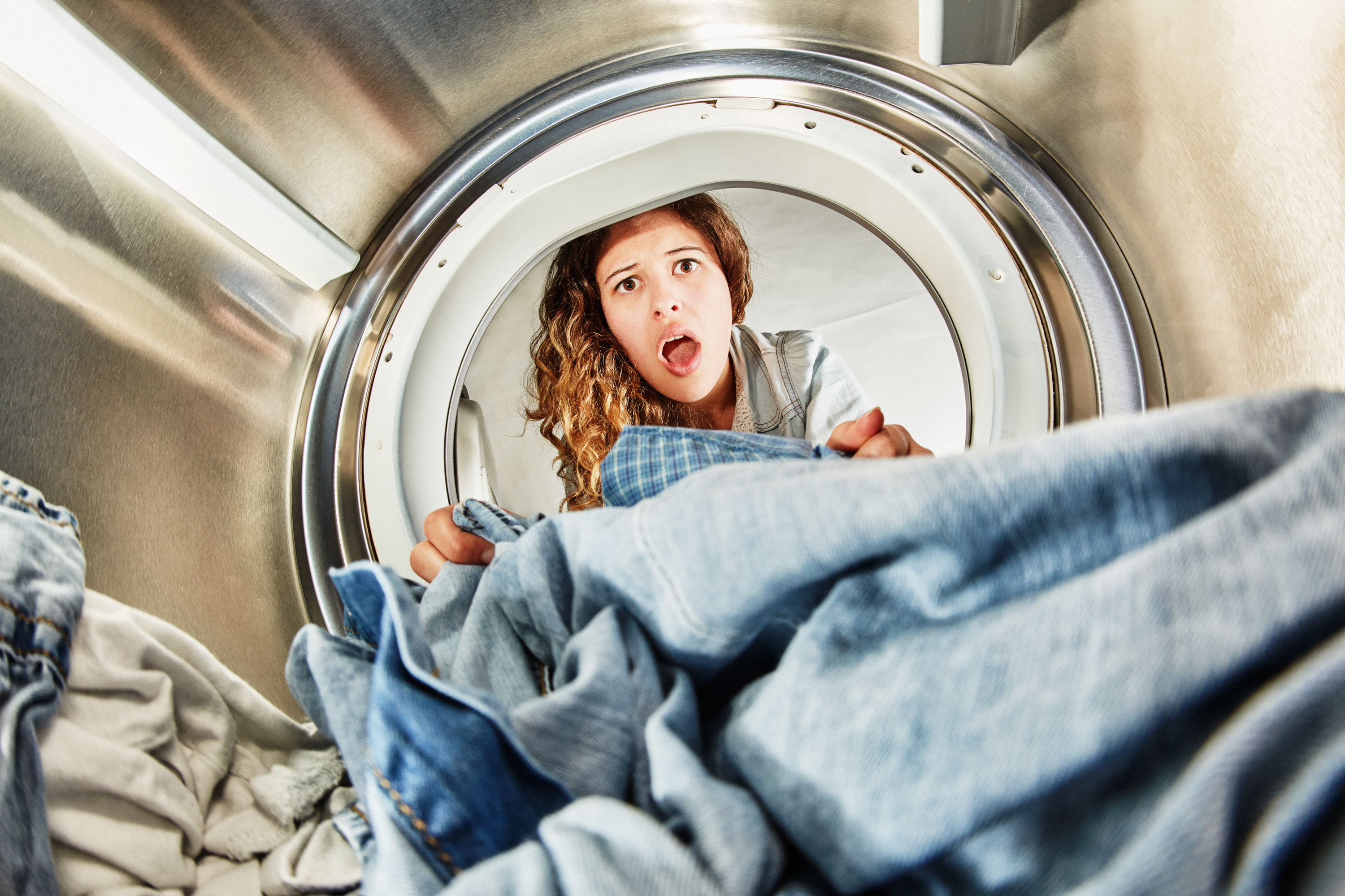 young woman with curly hair gasps open-mouthed as she checks out her newly dried clothes. Unusual angle from inside the appliance itself.