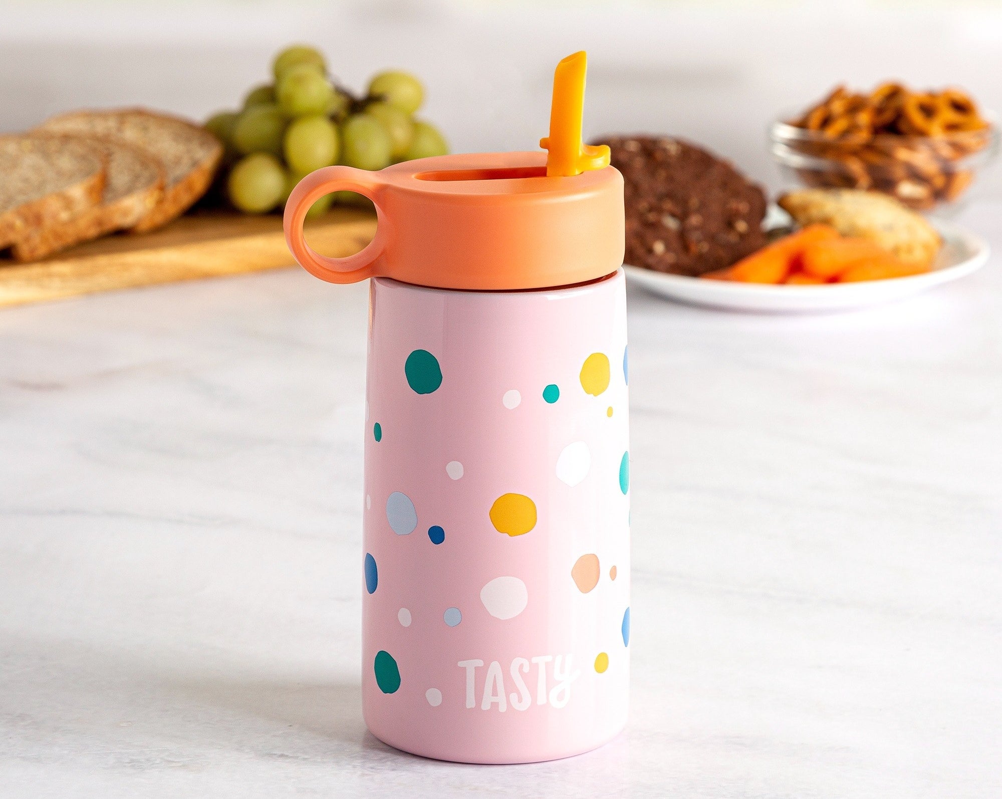 A pink and polka dot water bottle sitting on a counter with food in the background.