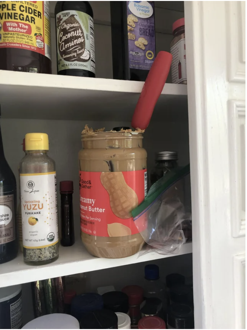 An open jar of peanut butter with a utensil sticking out of it on a cupboard shelf