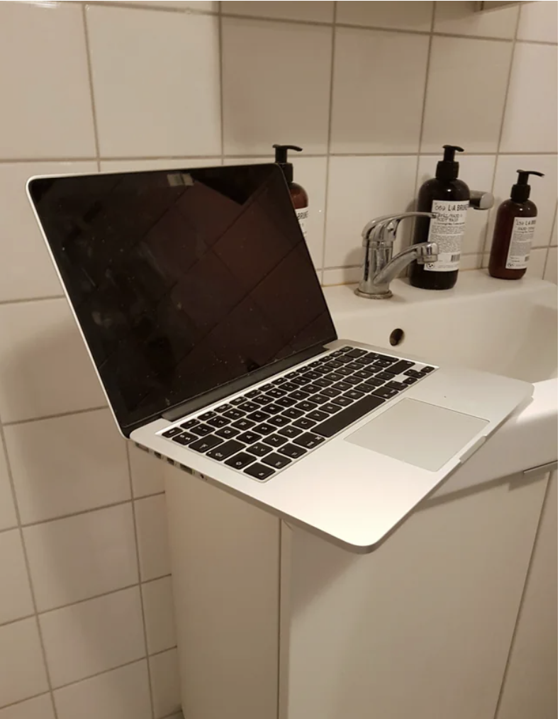 An open laptop perched precariously on the edge of a small bathroom sink