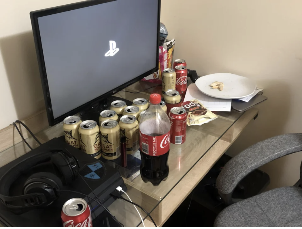 A computer screen and game console on a table alongside multiple beverage cans and bottles, snacks, and a plate