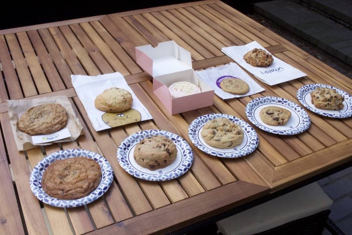 Four cookies are arranged on bakery bags; another one is in an open bakery box. Five more cookies are on paper plates. Most of the cookies are large and have visible chocolate chips.