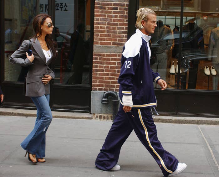 Victoria and David Beckham walking down the street. David is wearing a baggy tracksuit and Victoria is wearing a blazer, jeans and heels