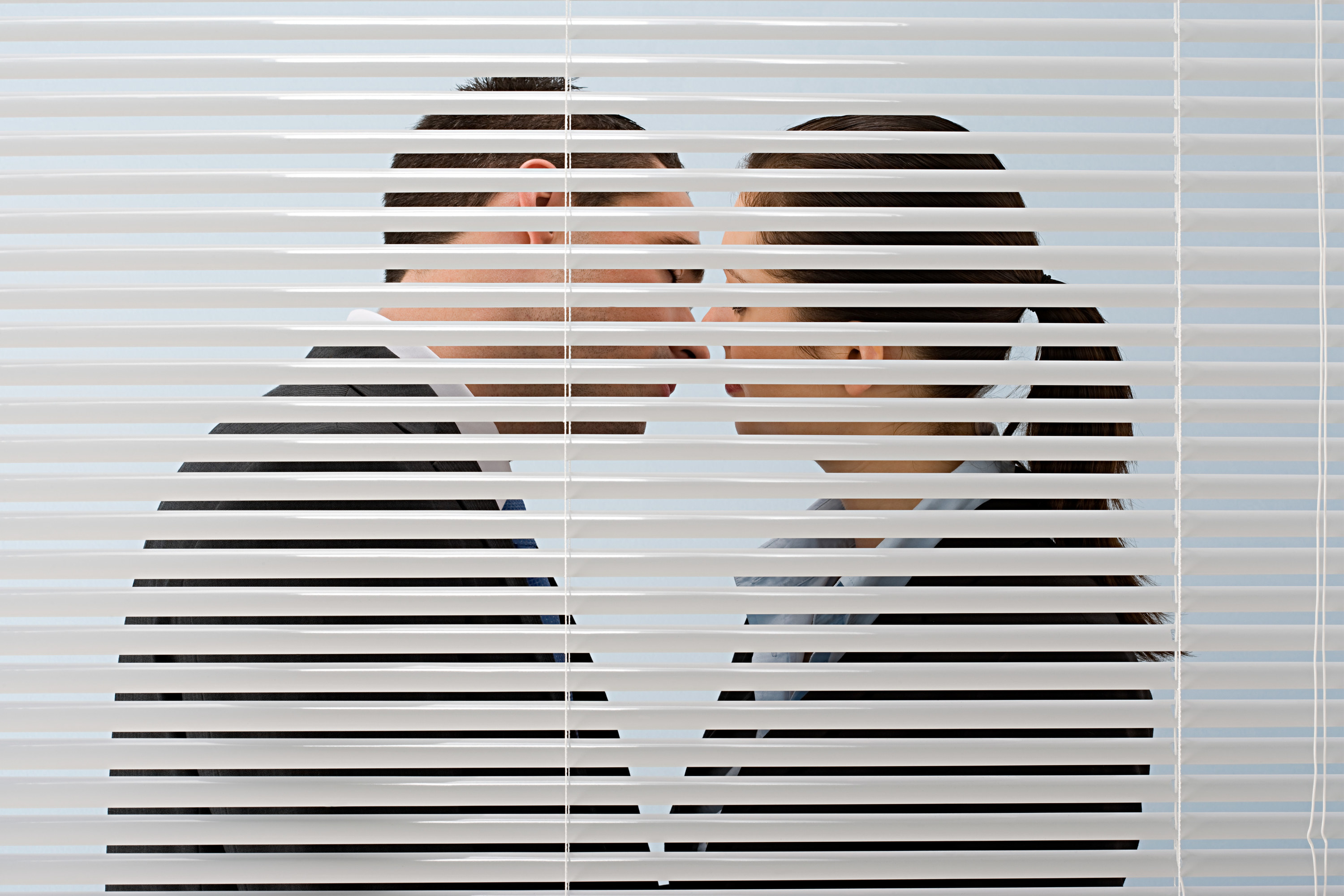 Two coworkers kiss behind the blinds of an office room