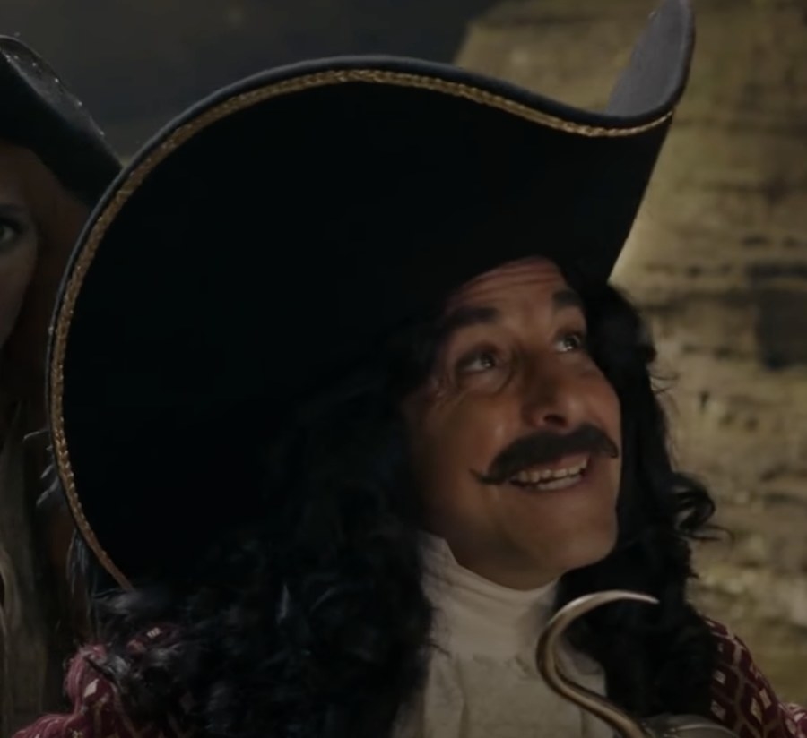 actor Stanley Tucci smiles in a mustache, long black wig, and pirate hat, with a hook under his chin
