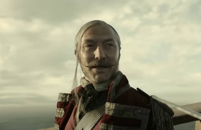 actor Jude Law has a mustache and long white hair and stands in a pirate coat in front of a cloudy sky
