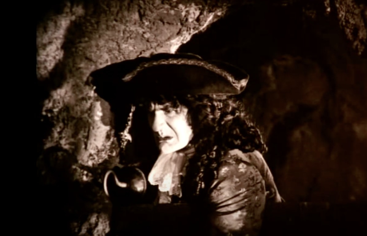 in black and white, a man in a pirate hat with a hook for a hand frowns while leaning against a stone wall