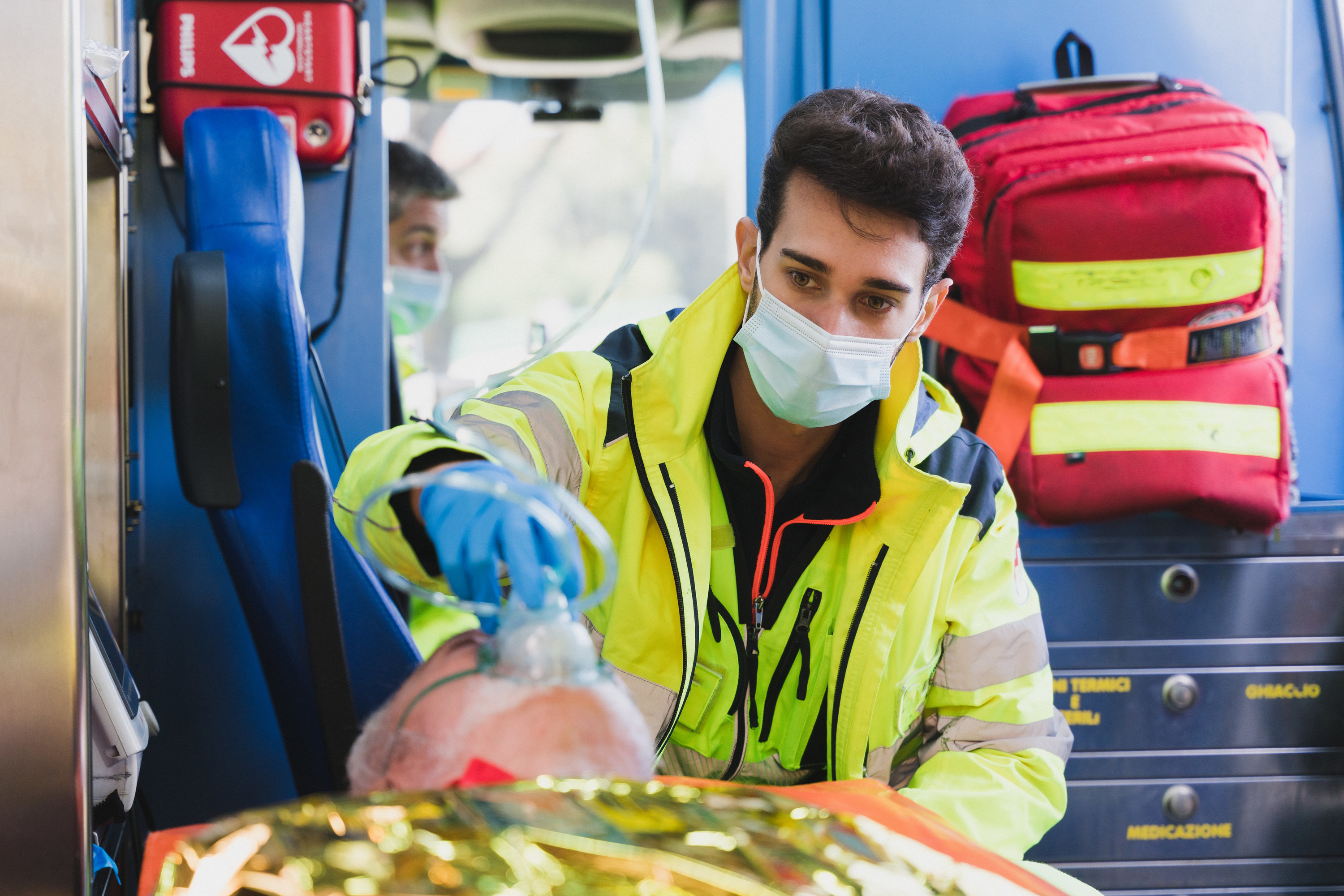 elder man in a ambulance wearing oxygen mask and lying on a stretcher unconscious with a paramedic taking care of him