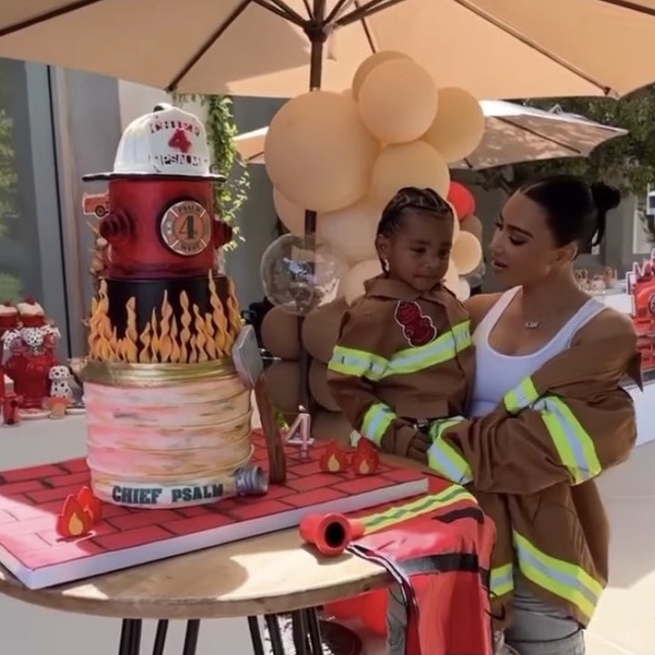 Kim holds Psalm next to a tiered cake that has a fire fighters hat on top and flames