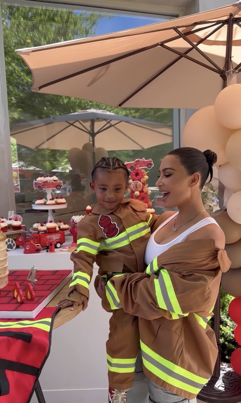 Kim holds Psalm in front of a table of cupcakes. They are both wearing fire fighters jackets