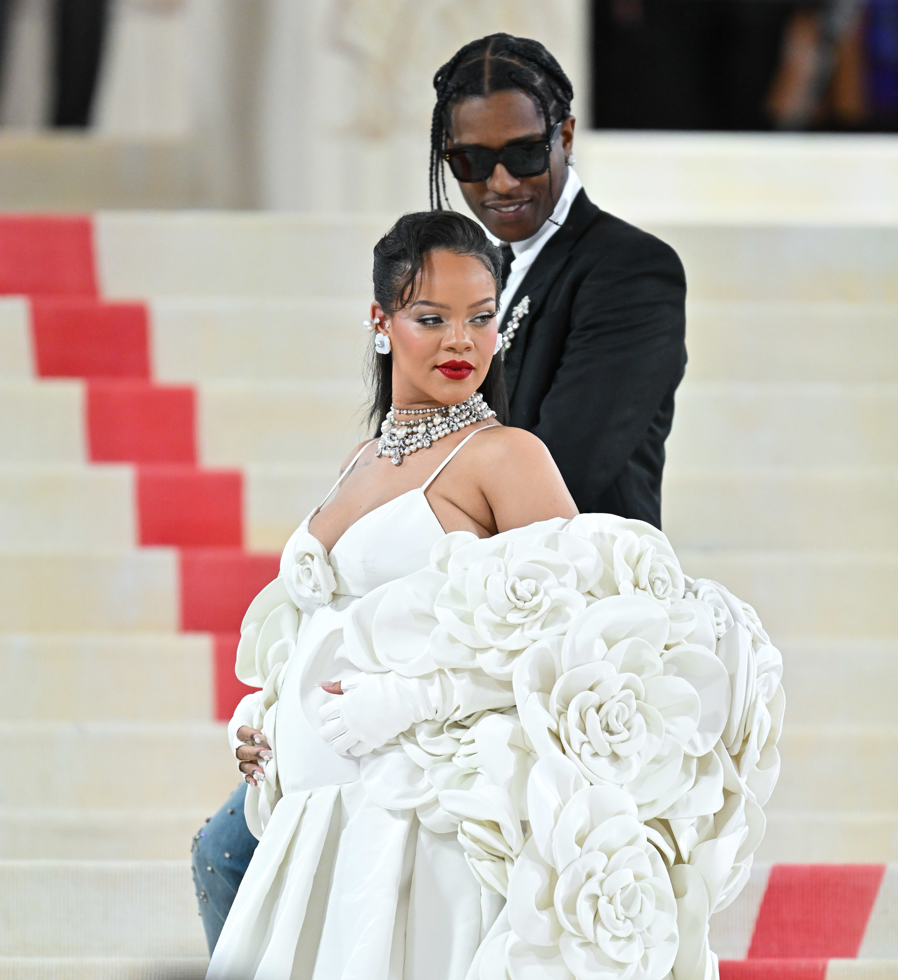 Rhianna poses in an extravagant evening gown on the steps of The Met prior to The Met Gala with A$AP Rocky standing behind peering down toward Rhianna from a few steps above
