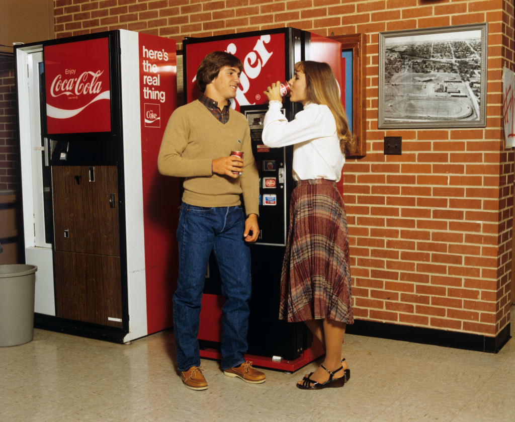 Two people drinking soda in front of the soda machine