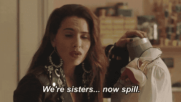 A woman looks to someone off-camera and says 'We're sisters&hellip; now spill.&quot;