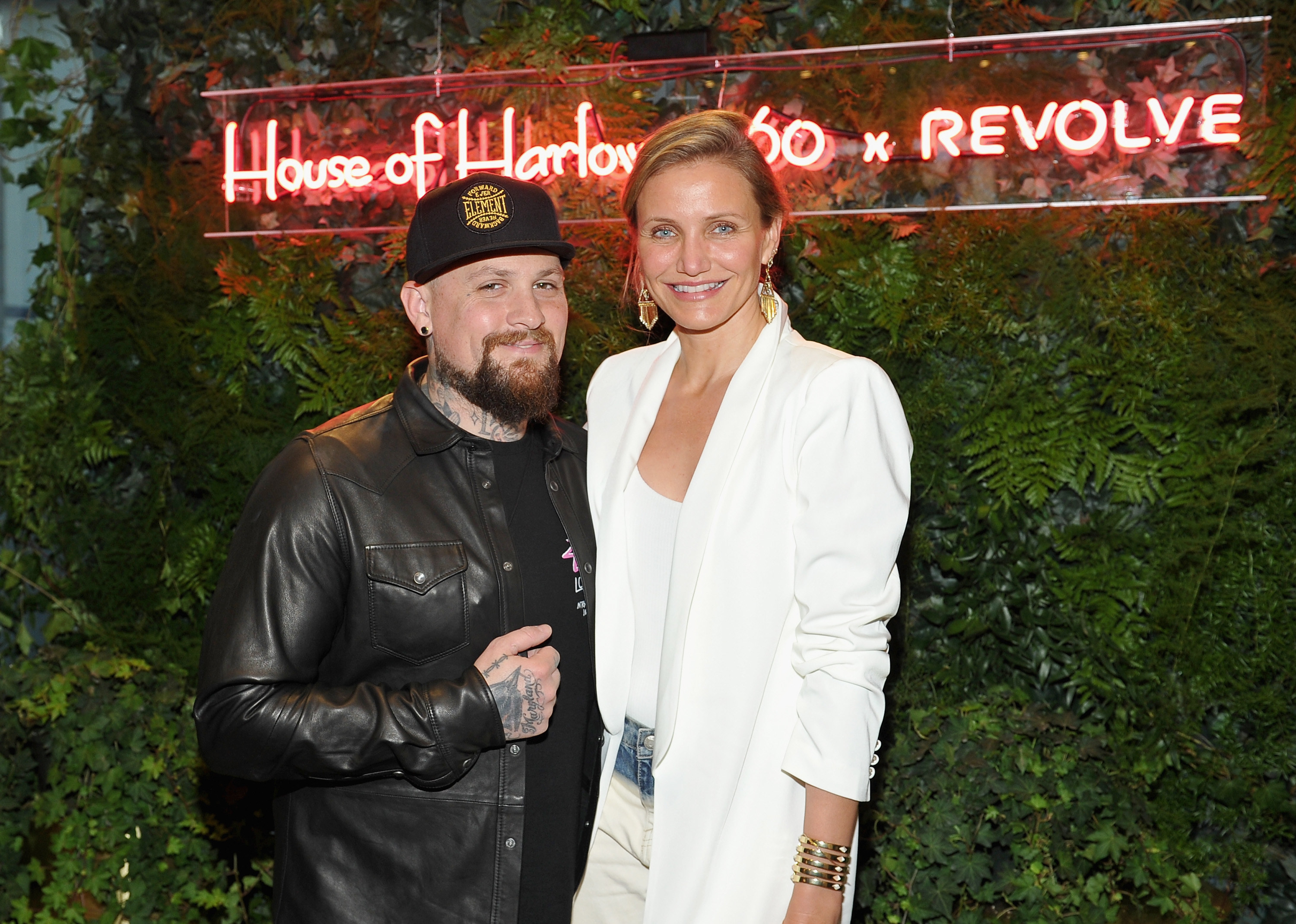 Benji Madden poses with his arm around wife Cameron Diaz in front of a backdrop with greenery and a neon sign