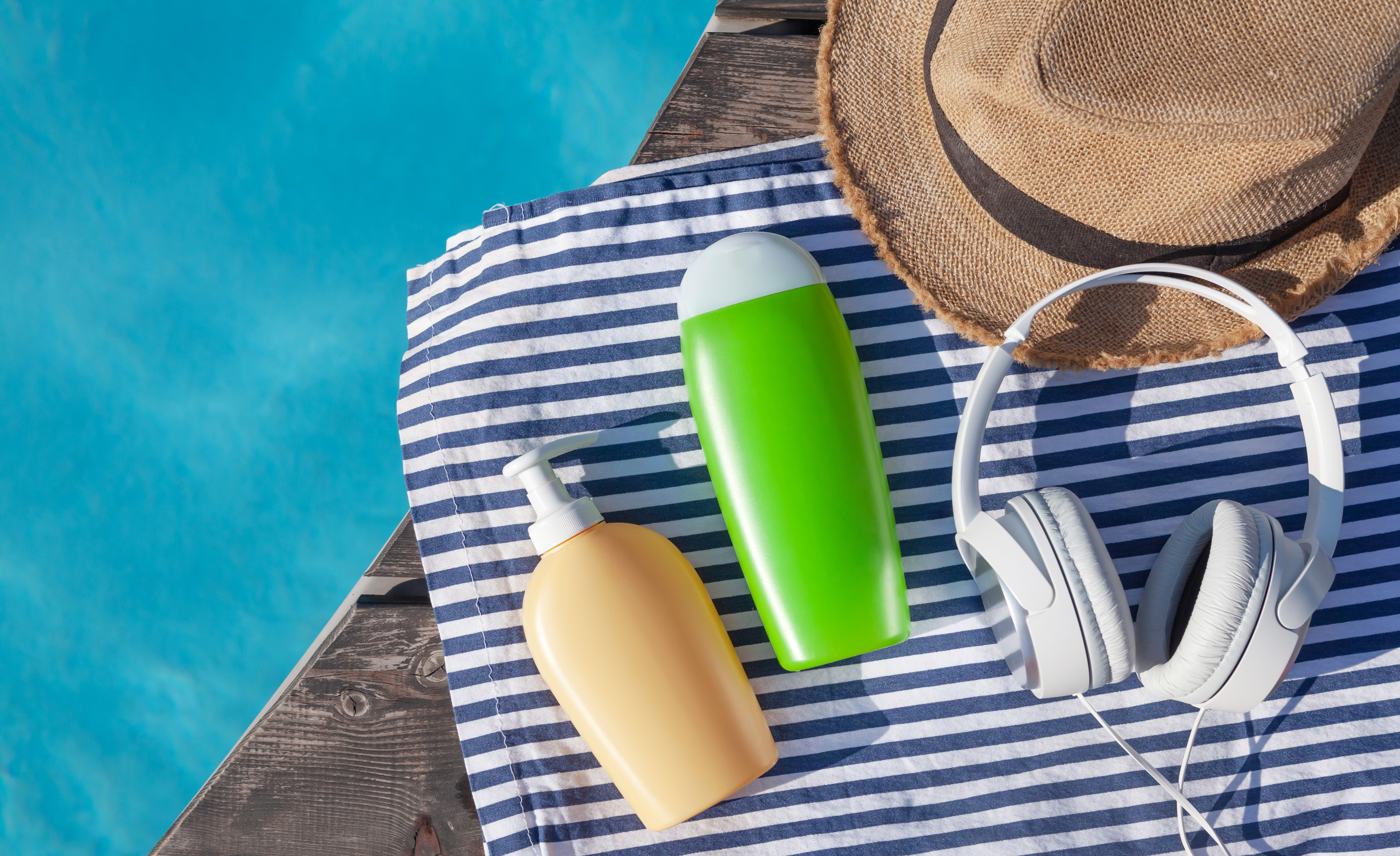 Two bottles of sunscreen on a striped towel poolside next to headphones and a sun hat