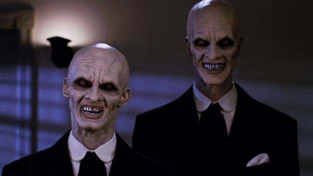 An image of two humanoid monsters in black suits with black shirts. They are bald, have deepset eyes with lots of black shadow around, dark veins and deep lines on their faces, and pointy teeth.